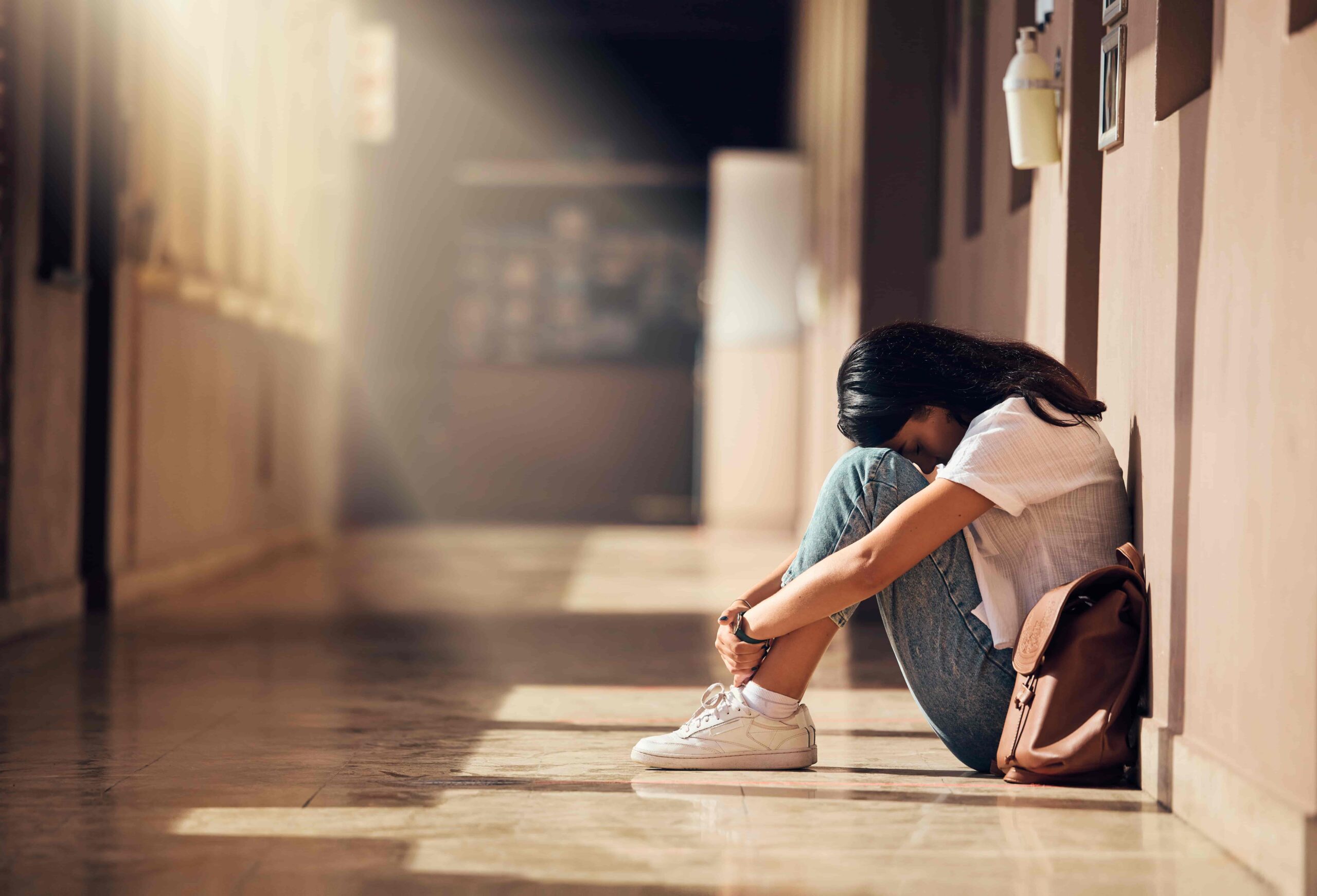 A teenage girl sits alone in a school hallway with her head down and knees pulled close, appearing sad and overwhelmed.