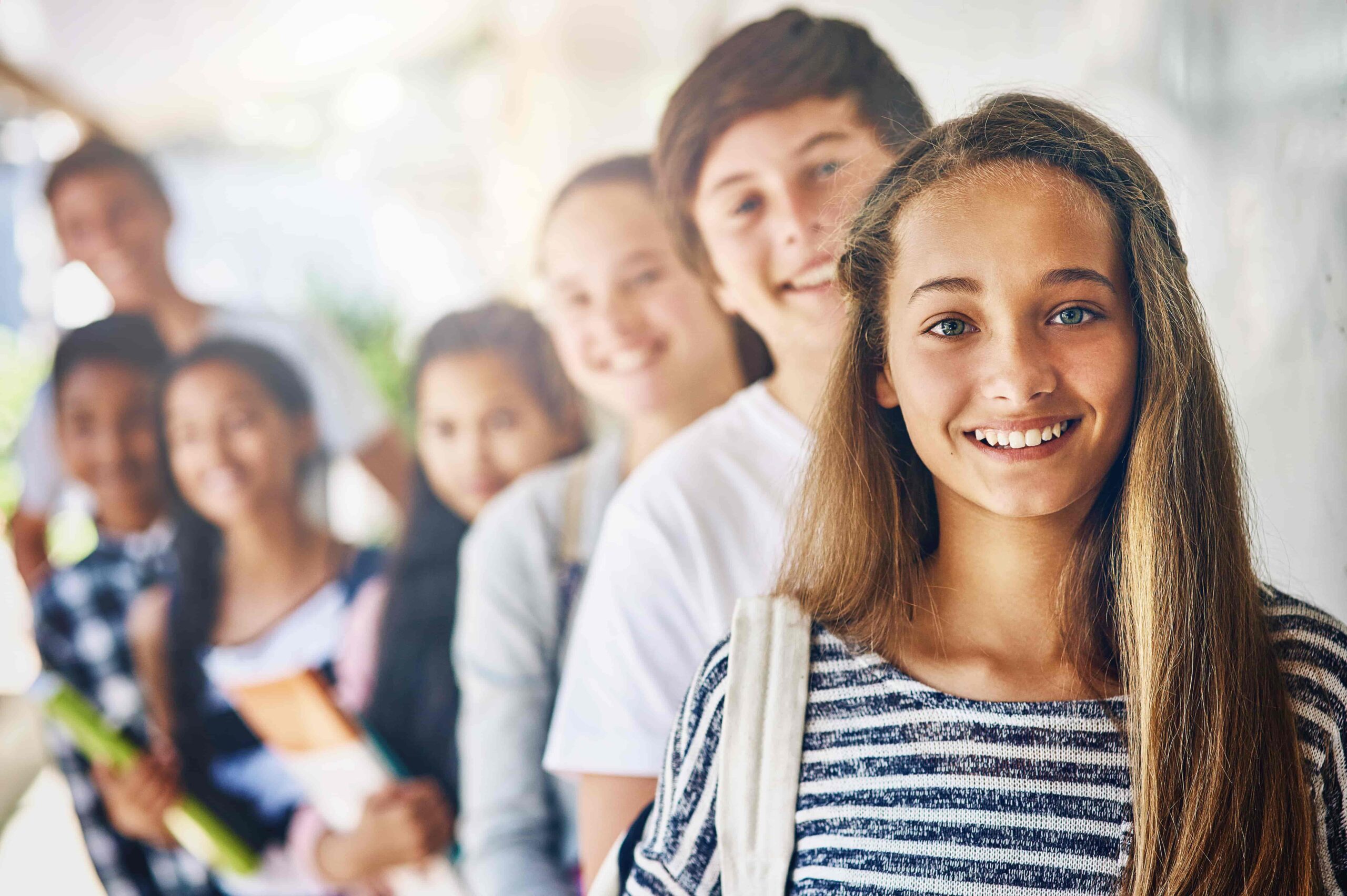 A smiling teenage girl stands in front of a group of friends in a school hallway, looking confident and relaxed.
